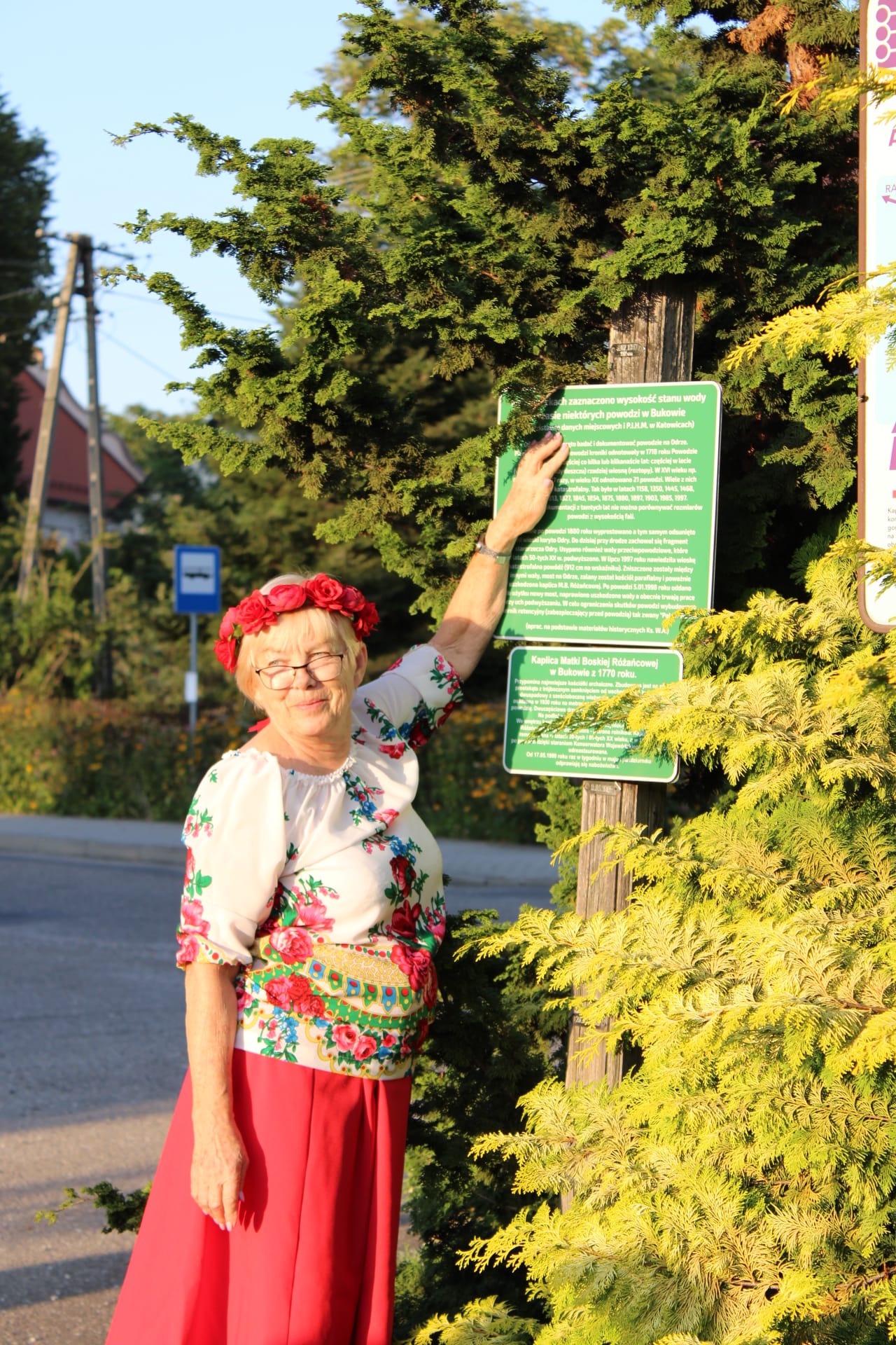 Magdalena Nieszporek z KGW Buków przy drewnianym elemencie, na którym umieszczone są tabliczki z datą powodzi, umiejscowione na takiej wysokości, do której sięgała woda w sołectwie. Fot. Zofia Błaszczok.